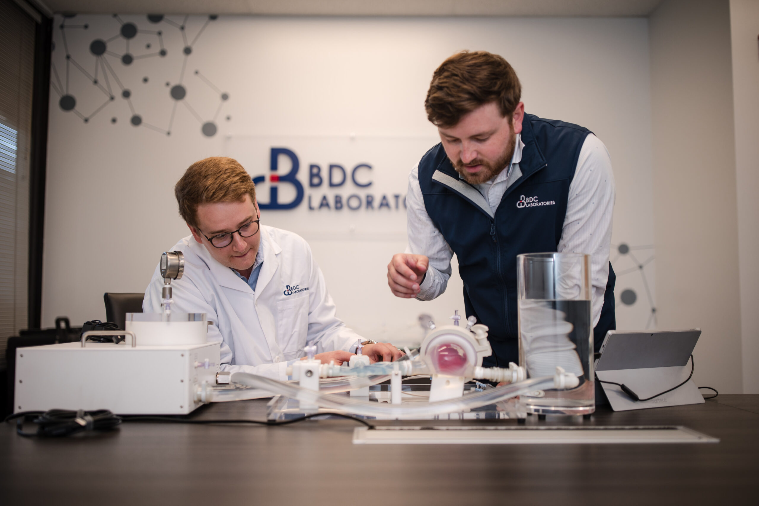 Two people in lab coats collaborate over scientific equipment at a table, with a video documenting their discoveries in the laboratory setting.