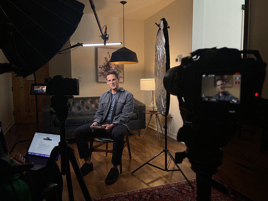 A man sits on a chair in front of a couch, being filmed by cameras and surrounded by lighting equipment in a home studio setup during a content planning call.