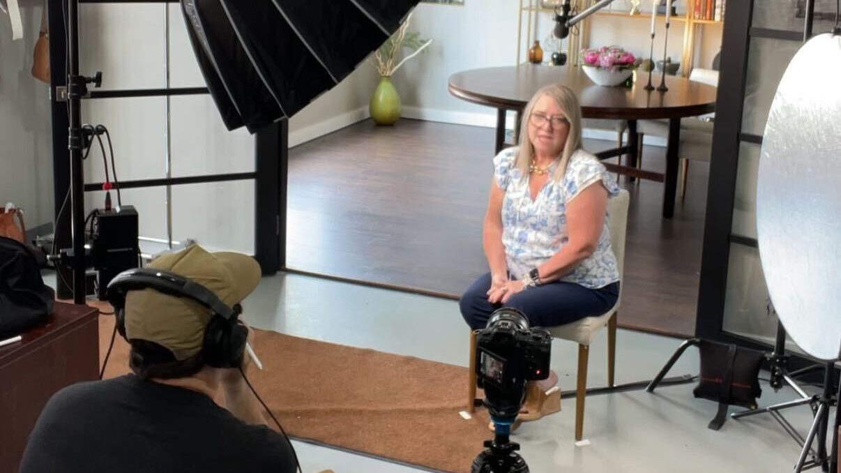 A woman sits on a chair facing a camera and lighting equipment in a home setting while a person with headphones conducts a Content Planning Call interview.