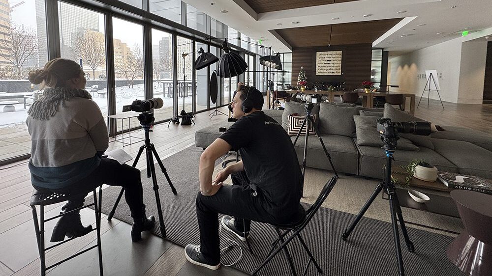 A woman is interviewed on camera by a man wearing headphones in a modern, well-lit room with large windows and photography equipment set up.
