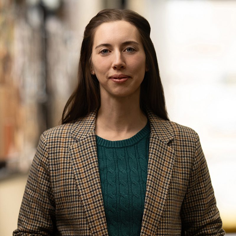A woman with long brown hair wearing a plaid blazer and green sweater stands indoors, looking at the camera with a neutral expression—perfect for a brand audit or sign up campaign visual.